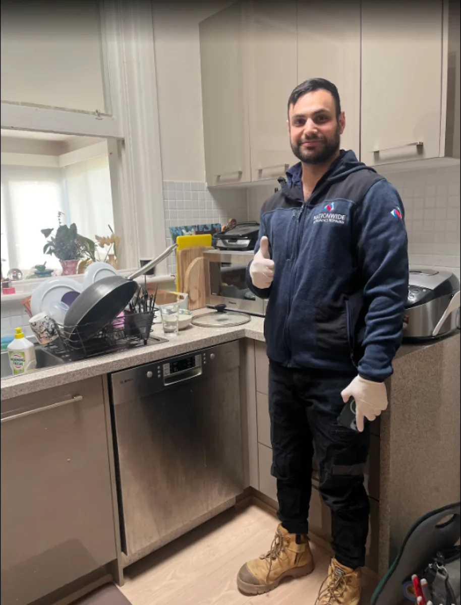A NAR technician standing in front of a repaired Bosch dishwasher in a client’s home, giving a thumb up.